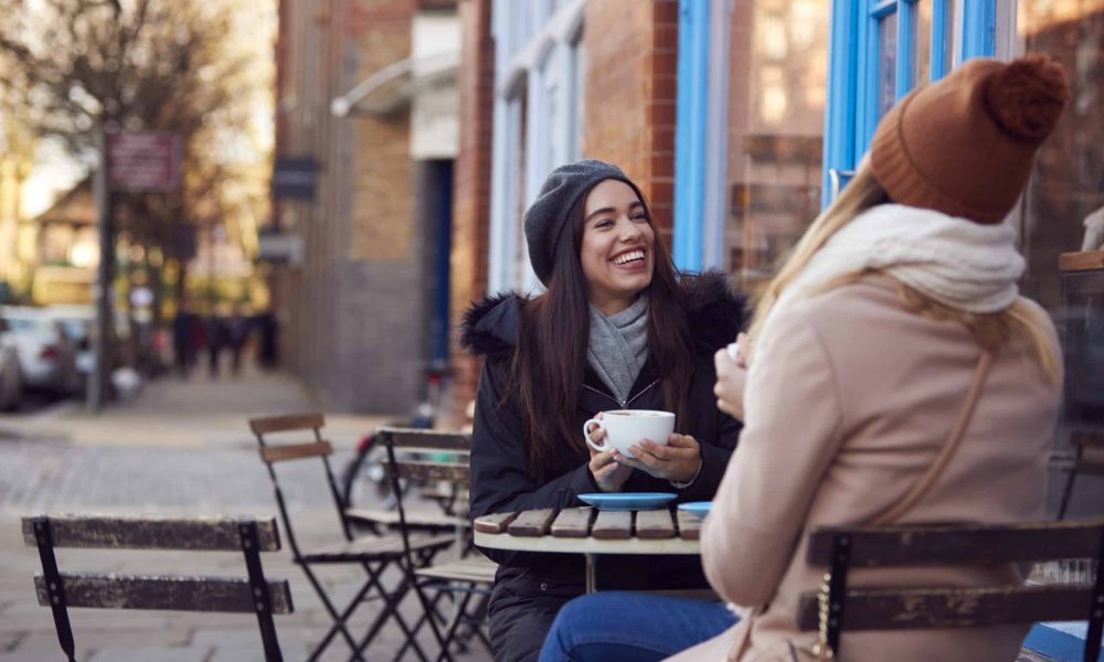 Two Female Friends Meeting Sitting Outside Coffee Shop On City High Street